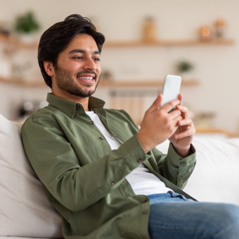 a young man sitting on a sofa smiles while looking at his phone.