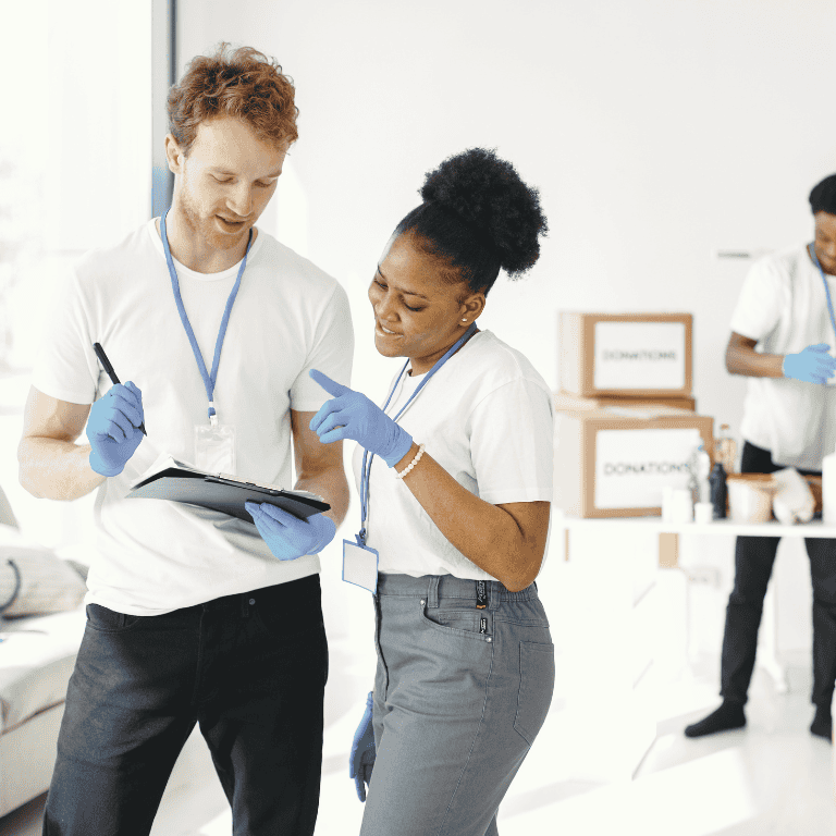Two volunteers, a man and a woman, wearing white t-shirts, blue gloves, and ID lanyards, collaborate in a bright room while reviewing notes on a clipboard. In the background, another volunteer works near stacked cardboard boxes labeled "Donations."
